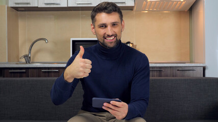 Bearded smiling man gamer plays game on smartphone wins rejoices in victory shows thumb up like. Male in blue sweater sits on couch at home holds smartphone in hands on background kitchen - Powered by Adobe