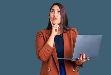 Young beautiful brunette woman using laptop serious face thinking about question with hand on chin,...