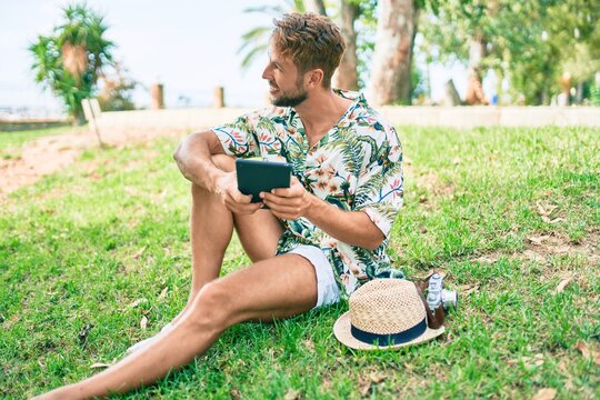 Caucasian handsome man smiling happy using touchpad device sitting on the grass at the park
