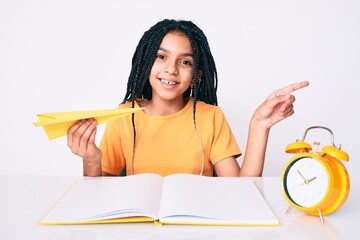 Young african american girl child with braids holding paper airplane while studying smiling happy pointing with hand and finger to the side