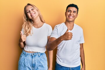 Young interracial couple wearing casual white tshirt doing happy thumbs up gesture with hand. approving expression looking at the camera showing success.