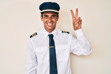 Young hispanic man wearing airplane pilot uniform showing and pointing up with fingers number two while smiling confident and happy.