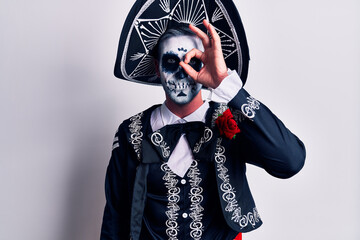 Young man wearing mexican day of the dead costume over white doing ok gesture with hand smiling, eye looking through fingers with happy face.