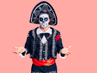 Young man wearing day of the dead costume over background clueless and confused expression with arms and hands raised. doubt concept.