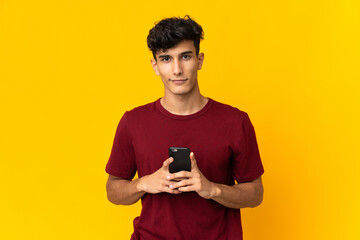 Young Argentinian man isolated on yellow background looking at the camera and smiling while using the mobile