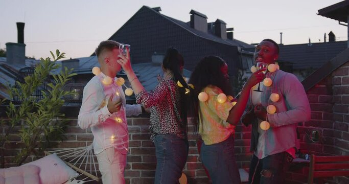 Attractive friendly smiling young mixed race company dancing with glasses of wine and yellow lighted bulbs on summer party on terrace at sundown