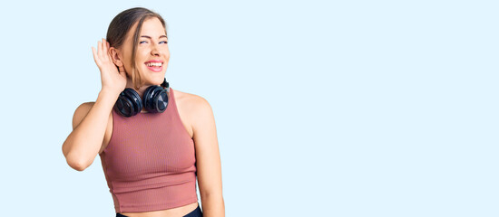 Beautiful caucasian young woman wearing gym clothes and using headphones smiling with hand over ear listening an hearing to rumor or gossip. deafness concept.