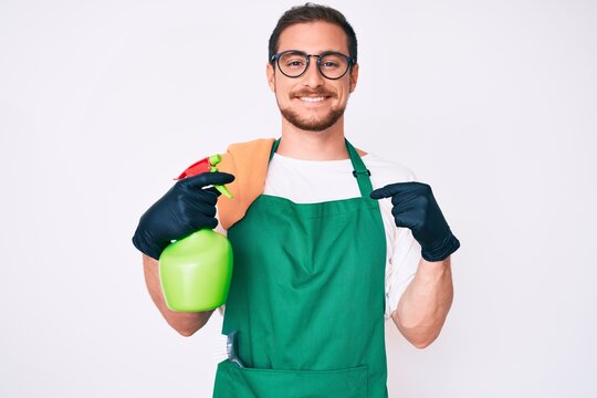Young handsome man wearing apron holding sprayer pointing finger to one self smiling happy and proud