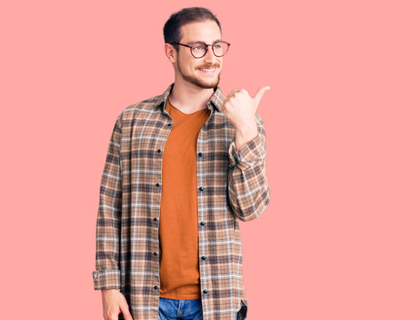 Young handsome caucasian man wearing casual clothes and glasses smiling with happy face looking and pointing to the side with thumb up.