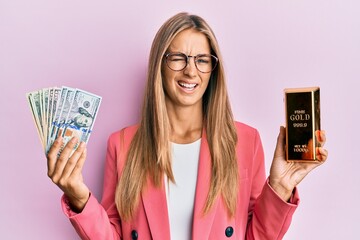Young blonde woman wearing business style holding gold ingot and dollars winking looking at the...