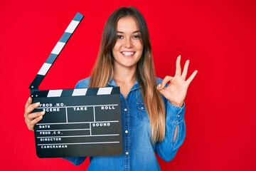 Beautiful caucasian woman holding video film clapboard doing ok sign with fingers, smiling friendly...