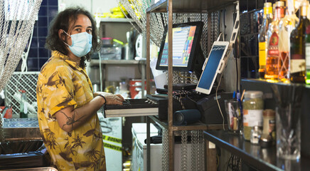 Waiter counting money from the cash register inside a bar. He wears a face mask. 