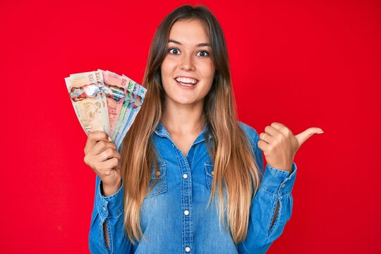 Beautiful caucasian woman holding canadian dollars pointing thumb up to the side smiling happy with open mouth
