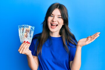 Young beautiful caucasian girl holding 100 swiss franc banknotes celebrating achievement with happy smile and winner expression with raised hand