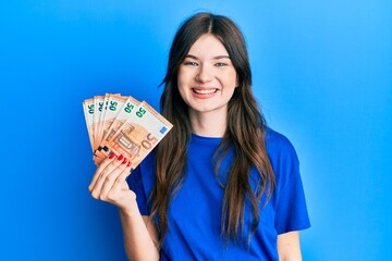 Young beautiful caucasian girl holding bunch of 50 euro banknotes looking positive and happy standing and smiling with a confident smile showing teeth