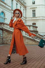 Outdoor full body autumn portrait of elegant woman wearing long orange trench coat, hat, sunglasses, cowboy ankle boots, with green croco textured bag, posing in street of European city