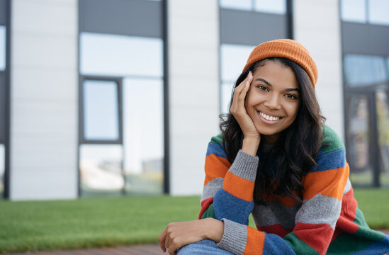 Portrait of beautiful smiling African American woman posing for pictures. Happy hipster girl wearing orange hat and colorful clothing sitting outdoors