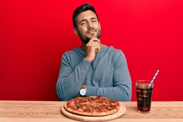 Handsome hispanic man eating tasty pepperoni pizza smiling looking confident at the camera with crossed arms and hand on chin. thinking positive.