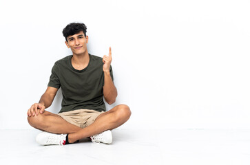 Young Argentinian man sitting on the floor showing and lifting a finger in sign of the best