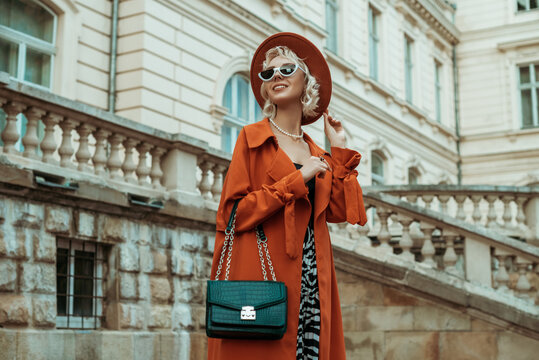 Outdoor autumn portrait of happy smiling woman wearing long orange trench coat, hat, sunglasses, with green croco textured bag, posing in street of European city. Copy, empty space for text