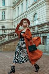 Street style full-length photo of elegant woman wearing long orange trench coat, black silk top, hat, animal print midi skirt,  cowboy ankle boots, with green bag, walking in street of European city