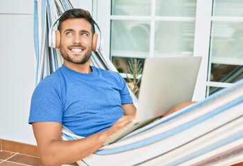 Young hispanic man relaxed working using laptop and headphones lying on the hammock at terrace.