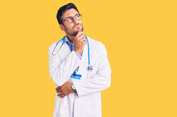 Young hispanic man wearing doctor uniform and stethoscope with hand on chin thinking about...