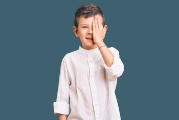 Cute blond kid wearing elegant shirt covering one eye with hand, confident smile on face and surprise emotion.