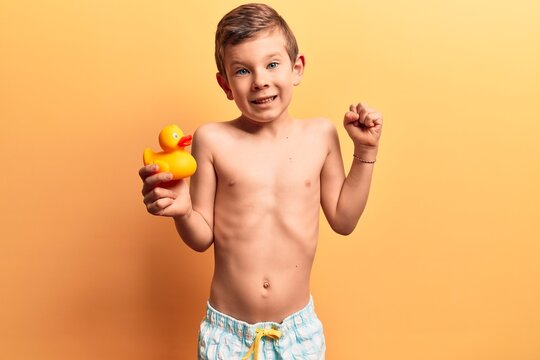Cute blond kid wearing swimwear holding duck toy screaming proud, celebrating victory and success very excited with raised arm