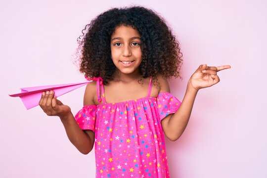 African american child with curly hair holding paper airplane smiling happy pointing with hand and finger to the side