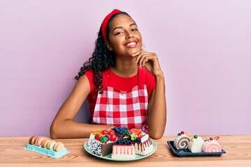 Young african american girl wearing baker uniform sitting on the table with sweets smiling looking confident at the camera with crossed arms and hand on chin. thinking positive.