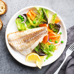 Fried fillet of sea white cod fish with juicy lettuce, capsicum, lemon, green peas on a large white dish on a gray background. Healthy balanced food. Top view,