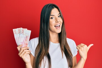 Young brunette woman holding 500 swedish krona banknotes pointing thumb up to the side smiling happy with open mouth