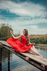 Vertical full length portrait of young woman sitting on the wooden pier near the river or lake touching clothes, dressed in red dress, looking away, copy space and nature blur background.