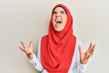 Young beautiful hispanic girl wearing traditional islamic hijab scarf crazy and mad shouting and yelling with aggressive expression and arms raised. frustration concept.