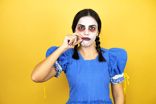 woman wearing a scary doll halloween costume over yellow background mouth and lips shut as zip with fingers. Secret and silent, taboo talking
