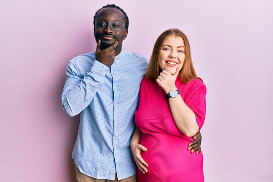 Young interracial couple expecting a baby, touching pregnant belly looking confident at the camera smiling with crossed arms and hand raised on chin. thinking positive.