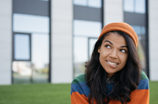Beautiful African American woman biting her lip, thinking, looking to copy space, standing outdoors. Close up portrait of young pensive hipster wearing casual clothes and orange hat 