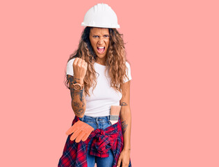 Young hispanic woman with tattoo wearing hardhat and builder clothes angry and mad raising fist frustrated and furious while shouting with anger. rage and aggressive concept.