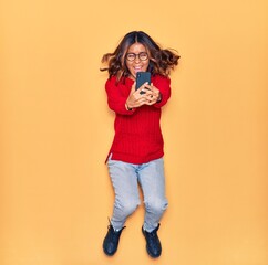 Young beautiful latin woman wearing glasses smiling happy. Jumping with smile on face make selfie by smartphone over isolated yellow background
