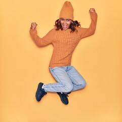 Fototapeta na wymiar Young beautiful latin woman wearing wool cap and sweater smiling happy. Jumping with smile on face celebrating with fists up over isolated yellow background