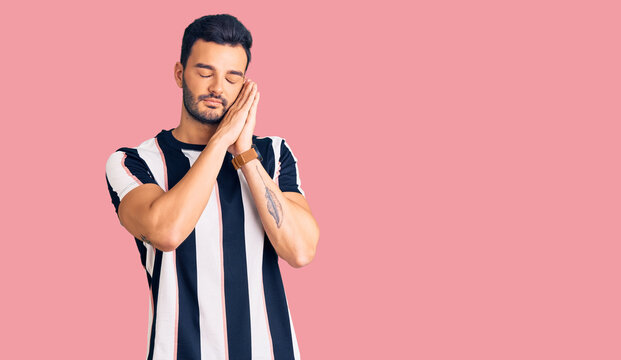 Young handsome hispanic man wearing striped tshirt sleeping tired dreaming and posing with hands together while smiling with closed eyes.