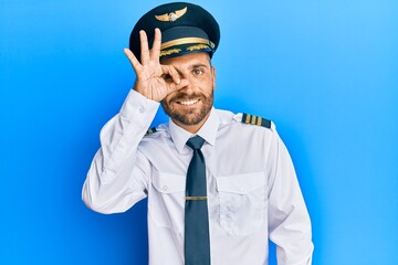Handsome man with beard wearing airplane pilot uniform doing ok gesture with hand smiling, eye looking through fingers with happy face.