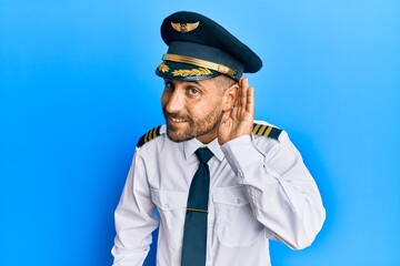 Handsome man with beard wearing airplane pilot uniform smiling with hand over ear listening an hearing to rumor or gossip. deafness concept.