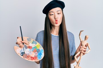 Young chinese woman wearing artist look with beret holding manikin making fish face with mouth and squinting eyes, crazy and comical.
