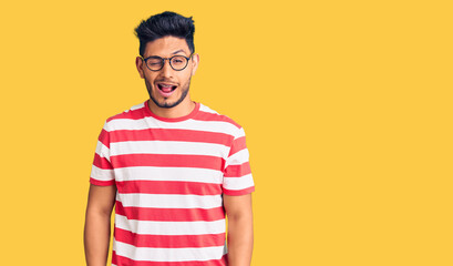 Handsome latin american young man wearing casual clothes and glasses winking looking at the camera with sexy expression, cheerful and happy face.