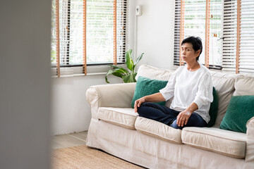 Senior old Asian woman sitting on sofa practicing yoga and meditation at home, retirement and wellbeing concept