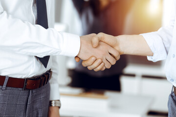 Two businessmen shaking hands in sunny office, close-up. Happy and excited business woman stands with raising hands at the background. Business people concept