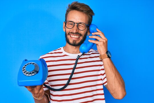 Handsome caucasian man with beard holding vintage telephone winking looking at the camera with sexy expression, cheerful and happy face.