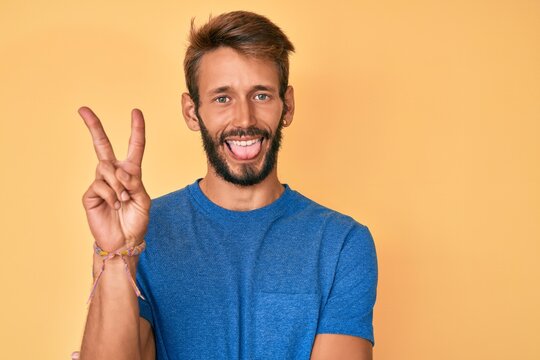Handsome caucasian man with beard wearing casual clothes smiling with happy face winking at the camera doing victory sign. number two.